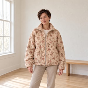 May include: A beige zip-up jacket featuring a repeating floral pattern in shades of muted red and brown. The jacket has a high collar, long sleeves, and two front pockets. It is worn over a plain white shirt and light-colored trousers.