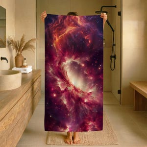 Könnte beinhalten: Ein rechteckiges Badetuch mit einem lebendigen Weltraum-Nebel-Druck, der wirbelnde Wolken in tiefem Magenta, Violett und leuchtendem Orange vor einem dunklen, sternenreichen Hintergrund zeigt. Das Design zeigt ein zentrales Leuchten.
