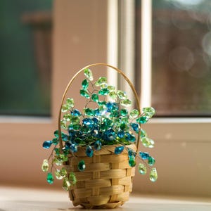 May include: A small woven light brown basket with a handle containing a decorative arrangement of wire branches adorned with numerous faceted crystal beads in various shades of lime green, emerald green, and deep ocean blue.