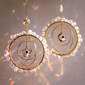 May include: Two circular gold-toned wire suncatchers hanging from chains. Each hoop is adorned with clear faceted beads. One features a central star charm with a small astronaut figure, while the other displays a crescent moon with an astronaut.