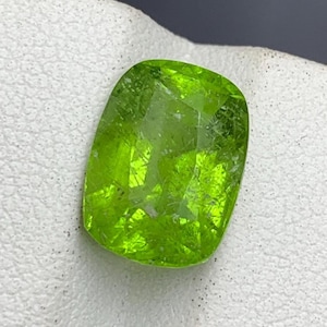 Könnte beinhalten: Ein kissenförmig geschliffener Peridot-Edelstein in leuchtendem Limettengrün mit facettierter Oberfläche. Der Stein weist interne Einschlüsse und einen hohen Grad an Transparenz auf und reflektiert das Licht auf seiner polierten Oberfläche.