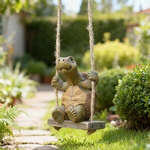 Può includere: Una statuetta di tartaruga marrone e marrone chiaro seduta su una piccola altalena rettangolare con texture in legno, sospesa da due spesse corde di iuta color naturale. La tartaruga ha un guscio testurizzato e un'espressione sorridente.