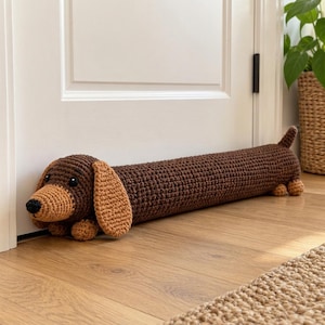 May include: A long, cylindrical crochet door draft stopper shaped like a dachshund. The body is dark brown, while the snout, ears, and small paws are a lighter tan color. It features black embroidered eyes and a black nose.