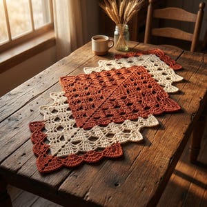 Può includere: Un runner da tavola rettangolare all'uncinetto con motivi quadrati alternati in filato color arancio bruciato e crema. Il design presenta intricati motivi traforati con bordi smerlati, creando un aspetto geometrico stratificato.