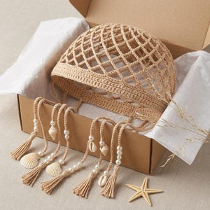 Op de afbeelding: Een beige gehaakte muts met een wijd netpatroon en een stevige rand. Verschillende bungelende gehaakte koorden hangen aan de rand, elk versierd met witte kunstparels, kleine natuurlijke schelpen en een kwastje aan het uiteinde van elk koord.