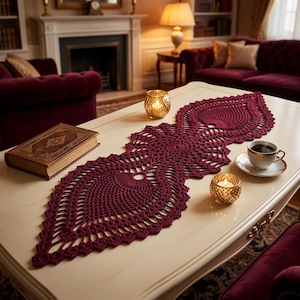 May include: A deep burgundy crochet table runner featuring an intricate, elongated floral-inspired pattern with openwork detailing. The runner is positioned centrally on a smooth, light-colored surface, showcasing a delicate, hand-crafted texture.