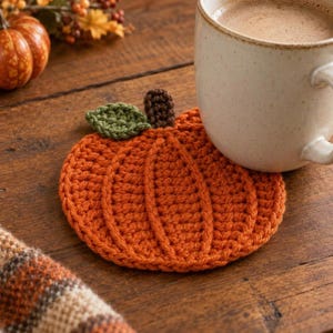 May include: A round, orange crochet coaster shaped like a pumpkin, featuring a small green leaf and a brown stem. The coaster is textured with vertical ridges and is designed to sit flat, providing a decorative base for a beverage mug.
