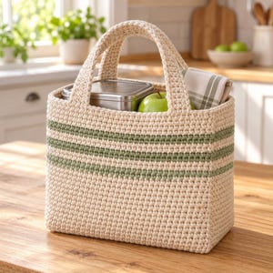 Könnte beinhalten: Eine rechteckige Häkeltasche aus cremefarbenem Garn mit zwei horizontalen salbeigrünen Streifen. Die Tasche hat zwei stabile Griffe und enthält einen rechteckigen metallischen Lebensmittelbehälter, einen grünen Apfel und ein gefaltetes weiß-grün kariertes Tuch.