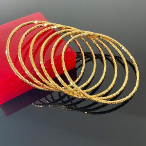 May include: A set of six thin, circular gold bangles with a textured, faceted surface. The bangles are arranged in a stacked, overlapping formation, reflecting light across their metallic surfaces to create a bright, shimmering appearance.