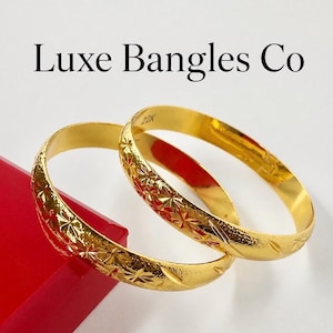 May include: Two gold bangles featuring a repeating star-like etched pattern on the exterior surface. The interior of one bangle is stamped with the text 22K. The text Luxe Bangles Co is displayed above the jewellery items.