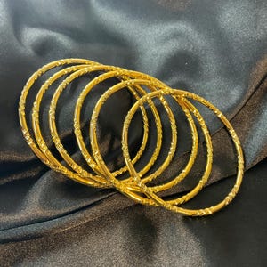 May include: A set of five thin, circular gold bangles, each featuring a repeating textured pattern of small, diagonal etched lines along the entire circumference of the metal bands, arranged in a slightly overlapping, cascading formation.