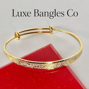 May include: A gold-toned expandable bangle bracelet featuring a textured, glittery finish on the lower half and smooth, polished metal on the upper half. The text Luxe Bangles Co is displayed above the bracelet in a black serif font.