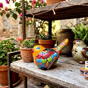May include: A colorful ceramic bird figurine featuring vibrant hand-painted floral patterns in red, yellow, blue, and green. The bird is positioned on a weathered wooden shelf next to a second, more neutral-toned ceramic bird and a stack of decorative tiles.