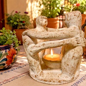 May include: A beige, textured candle holder featuring three stylized figures standing in a circle with arms linked, surrounding a central bowl that contains a small, lit yellow candle. The sculpture has a weathered, matte finish.