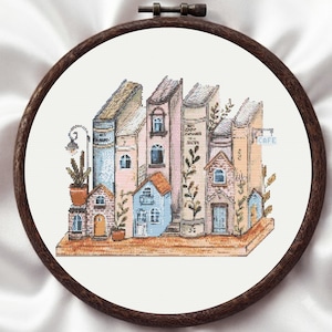 May include: A cross-stitch embroidery in a dark wood hoop featuring a row of books designed to look like a miniature street of buildings. The scene includes brick houses, a street lamp, potted plants, and a small sign with the text CAFE.