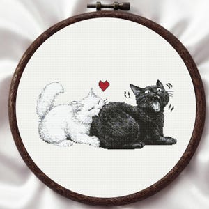 May include: A circular wooden embroidery hoop containing a cross-stitch pattern on white fabric. The design features a white cat sniffing a black cat, which has its mouth open in a surprised expression. A small red heart is stitched above the cats.
