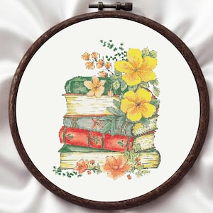 May include: A cross-stitch embroidery piece in a dark wood circular hoop featuring a stack of four books with green, red, and yellow spines. The books are decorated with clusters of bright yellow and orange flowers and green foliage.