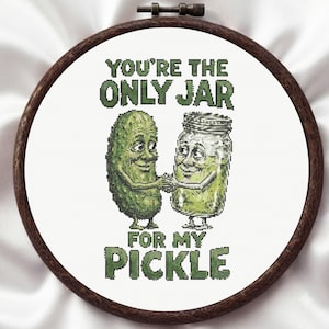 Op de afbeelding: Een ronde houten borduurring met witte stof en een kruissteekontwerp van een groene augurk en een glazen pot die elkaars hand vasthouden. De tekst YOU'RE THE ONLY JAR FOR MY PICKLE staat in groene blokletters boven en onder de figuren.