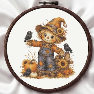 May include: A cross-stitch pattern in a dark wood embroidery hoop featuring a scarecrow wearing a hat and blue overalls, surrounded by sunflowers, pumpkins, and three black birds. The design uses warm autumn colors and detailed stitching.