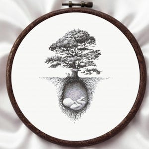 以下が含まれることがあります： 円形の木製刺繍枠に、黒いクロスステッチのデザインが施された白い布が張られています。地上には詳細な木が描かれており、その根が空洞を作り、その中に小さく丸まった赤ん坊が収まっている様子が表現されています。