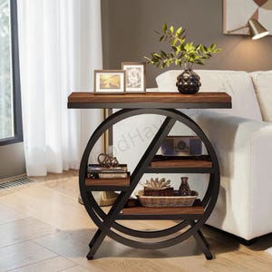 May include: A round black metal side table with a dark wood top and two interior shelves. The frame features a circular design with a diagonal support. The table holds two framed photos, a small plant in a black vase, books, a speaker, and a basket.