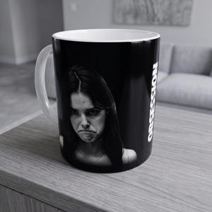 Op de afbeelding: Een zwarte keramische mok met een witte binnenkant en een wit handvat. De buitenkant toont een zwart-wit portretfoto van een persoon met een serieuze uitdrukking, vergezeld van de verticale witte tekst OBSESSION gedrukt op de zijkant.