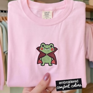 Op de afbeelding: Een lichtroze T-shirt met korte mouwen en een centrale geborduurde afbeelding van een groene kikker in een donkerrode vampiercape met een hoge kraag. De kikker heeft zichtbare witte hoektanden en rode wangen. Comfort Colors label in de nek.