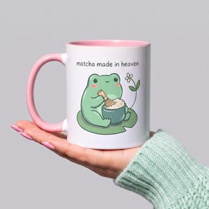 May include: A white ceramic mug with a light pink handle and interior rim. The mug features a graphic of a green frog sitting on a lily pad, whisking a bowl of matcha, with the text "matcha made in heaven" printed above the illustration.