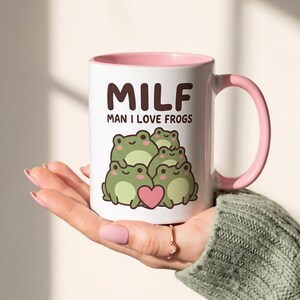 Puede incluir: Una taza de cerámica blanca con interior y asa de color rosa, que presenta un gráfico de cuatro ranas verdes y un corazón rosa. El texto "MILF" está impreso en letras grandes de color marrón oscuro sobre el texto "MAN I LOVE FROGS".