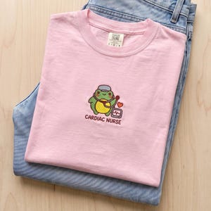 Könnte beinhalten: Ein hellrosa T-Shirt mit einer Stickerei, die einen grünen Frosch mit Krankenschwesterhaube und Stethoskop zeigt. Das Motiv enthält ein kleines rotes Herz, ein medizinisches Monitor-Symbol und den Text "CARDIAC NURSE" in dunkelbrauner Schrift.