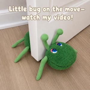 May include: A green plush bug doorstop with two antennae and large blue eyes, positioned under a white door. The text "Little bug on the move- watch my video!" is displayed above the bug. The bug has a textured fabric surface and limbs.