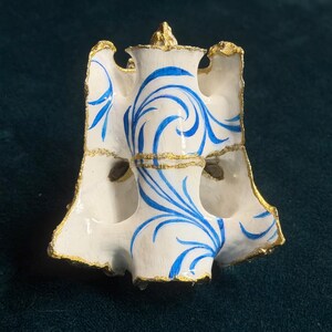 May include: A white ceramic vertebra sculpture featuring hand-painted blue swirling patterns and textured gold leaf detailing along the edges. The bone-shaped object has a hollow centre and a flared, irregular structure with a smooth finish.