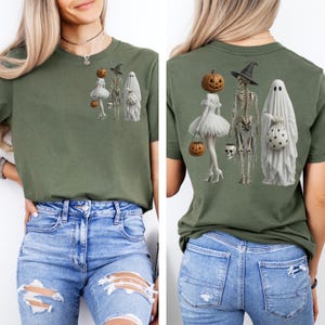 May include: An olive green t-shirt featuring a graphic print of a pumpkin-headed figure in a white dress, a skeleton in a witch hat, and a ghost holding a star-patterned bag. The same graphic appears in a smaller size on the front chest.