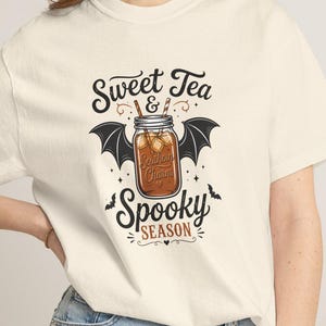 Peut inclure: Un t-shirt à manches courtes de couleur crème avec un motif représentant un bocal en verre rempli de thé, entouré d'ailes de chauve-souris noires. Le texte indique Sweet Tea & Spooky Season, accompagné de petites silhouettes de chauves-souris.