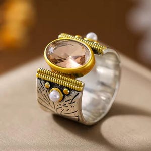 Peut inclure: Une large bague argentée ornée d'une pierre ovale à facettes de couleur pêche sertie dans un chaton doré. L'anneau présente des motifs de lignes gravées, des accents dorés texturés et deux petites perles sphériques blanches incrustées.