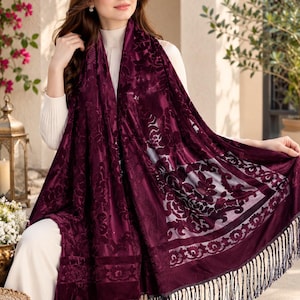 May include: A deep burgundy velvet scarf featuring a tonal floral burnout pattern, a rectangular shape, and long dark fringe trim along the bottom edge. The fabric displays a rich, textured appearance with a mix of opaque and sheer sections.