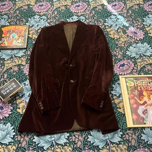 Puede incluir: Una americana de terciopelo marrón oscuro con solapa de muesca, dos botones frontales y tres botones en cada puño. La americana cuenta con un forro interior color canela y se muestra junto a un disco de Carnaby Street, una caja Good News y un póster.