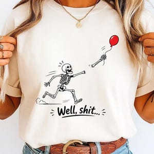May include: A cream-colored t-shirt featuring a black line-art graphic of a skeleton running after a detached hand holding a red balloon. Below the graphic, the text reads "Well, shit..." in a bold, black, sans-serif font with small decorative lines.