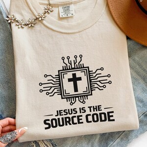 May include: An ivory t-shirt featuring a black graphic of a computer chip with a cross in the center and the text "JESUS IS THE SOURCE CODE" printed below. The shirt has a Comfort Colors label at the neckline and is paired with blue denim fabric.