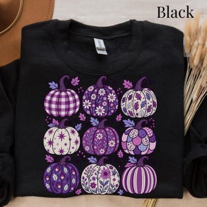 May include: A black crewneck sweatshirt featuring a graphic print of nine pumpkins in various shades of purple and white. Each pumpkin displays a unique pattern, including plaid, floral, star, leaf, and striped designs, arranged in a three-by-three grid.
