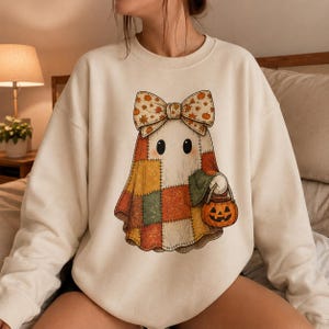 Könnte beinhalten: Ein cremefarbenes Sweatshirt mit Rundhalsausschnitt und einem zentralen Grafikdruck eines Geistes, der eine große Schleife mit Kürbis- und Blattmuster trägt. Der Geist ist in einen Patchwork-Stoff in Orange-, Gelb- und Grüntönen gehüllt.