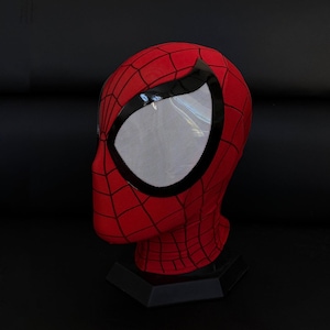 May include: A red fabric mask featuring a black web pattern across the surface. The mask includes large, almond-shaped eye lenses with a white mesh texture and thick, glossy black frames surrounding the perimeter of each eye opening.