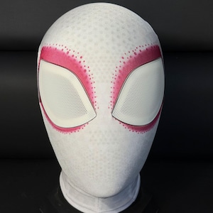 Könnte beinhalten: Eine weiße Vollkopfmaske mit einer strukturierten Stoffoberfläche und großen, mandelförmigen Augenlinsen. Die Augenöffnungen sind mit einem leuchtend pinkfarbenen, gepunkteten Airbrush-Muster umrandet, das in das weiße Material übergeht.