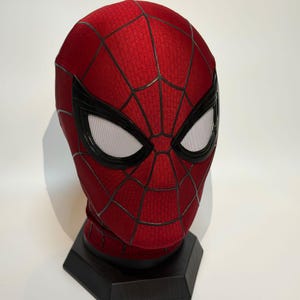 May include: A red fabric mask featuring a black web pattern across the surface. The mask includes two large, almond-shaped eye lenses with white mesh inserts framed by thick black borders, designed to cover the entire head and face area.