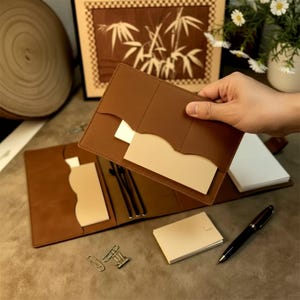 Peut inclure: Une couverture de carnet en cuir beige avec plusieurs poches et fentes intérieures, contenant des inserts en papier crème. L'ensemble comprend un petit bloc-notes assorti, un stylo à bille noir et deux trombones en métal argenté.