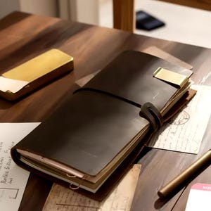 May include: A dark brown leather journal with a wrap-around strap closure, featuring a gold-toned metal clip on the top edge. The journal contains multiple pages and is accompanied by a gold-toned rectangular case and a gold-toned pen.