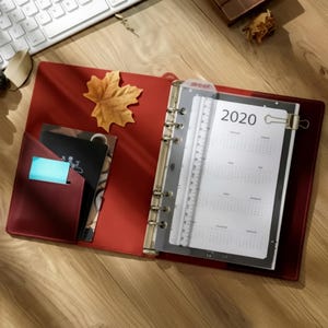 Può includere: Un raccoglitore aperto in pelle rossa con un meccanismo metallico a sei anelli, contenente una pagina di calendario 2020 e un righello in plastica trasparente da 15 cm. Il lato sinistro presenta una tasca con una carta nera e una linguetta blu.