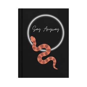 May include: A black hardback journal featuring a graphic of a coral-coloured snake with a geometric pattern. Above the snake is a white glowing circular ring and the white cursive text "Say Anyway" centred on the front cover of the notebook.