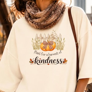 May include: An off-white short-sleeve t-shirt featuring a graphic of a pumpkin with a plaid bow in front of a white picket fence, surrounded by wheat stalks. The text reads, Plant for a harvest of kindness, with small acorns and leaves.