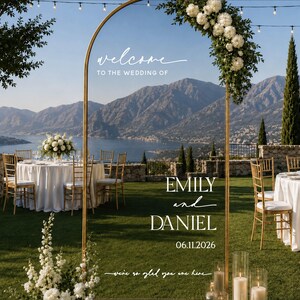 May include: A tall, arched gold metal wedding welcome sign featuring white text that reads: welcome TO THE WEDDING OF EMILY and DANIEL 06.11.2026 we're so glad you are here. The sign is decorated with a cluster of white flowers and greenery at the top.